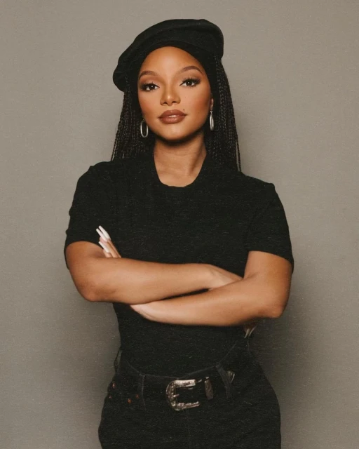 Portrait of a woman with braided hair wearing a beret, black top, and jeans, standing in front of a gray background with her arms crossed.