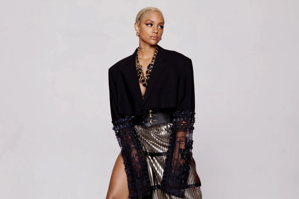 Woman in a black blazer, corset belt, gold skirt, and gold jewelry, looking to the side against a white background in a fashion editorial style.