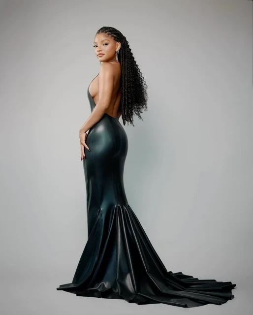 Woman in a black mermaid gown with a train, posing in a studio against a neutral background; the dress features a low back.