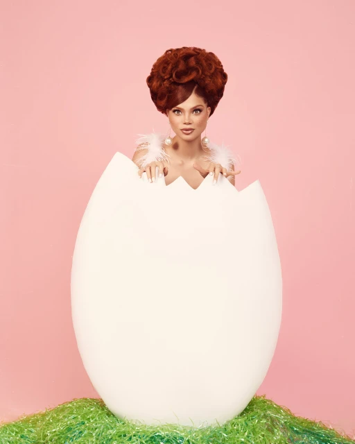 Adult woman emerges from a cracked eggshell in a playful studio shot, wearing white feathers and standing on a faux grass surface.