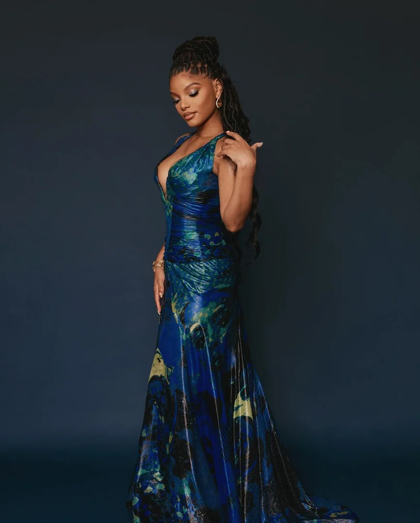 Halle Bailey wearing a long blue patterned evening gown with a plunging neckline against a dark studio background