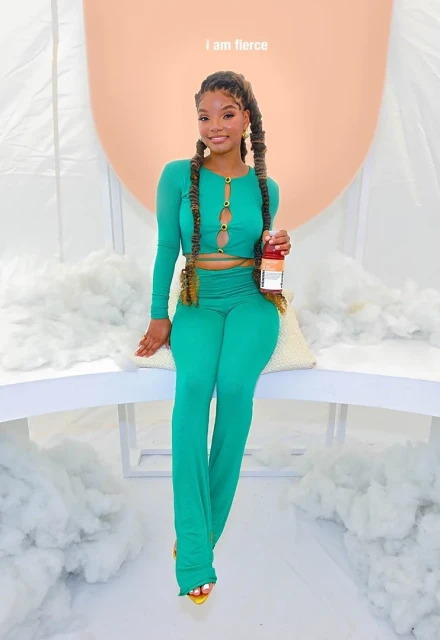 A woman with long braids, wearing a long sleeve green matching set, sits on a white ledge against a soft pink and white background.