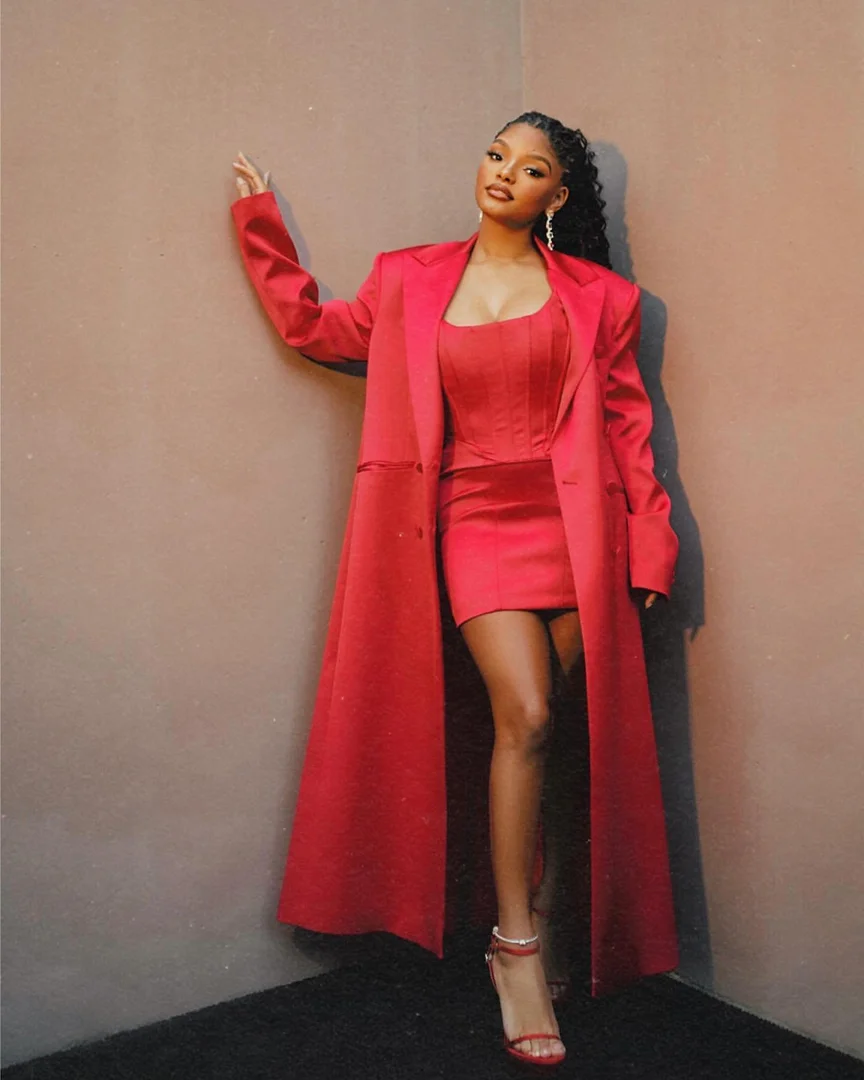 Woman in a red mini dress, jacket, and strappy heels poses in front of a grey corner, elegant and fashionable for an event.