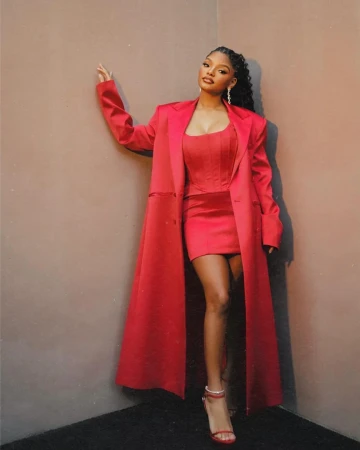 Woman in a red mini dress, jacket, and strappy heels poses in front of a grey corner, elegant and fashionable for an event.