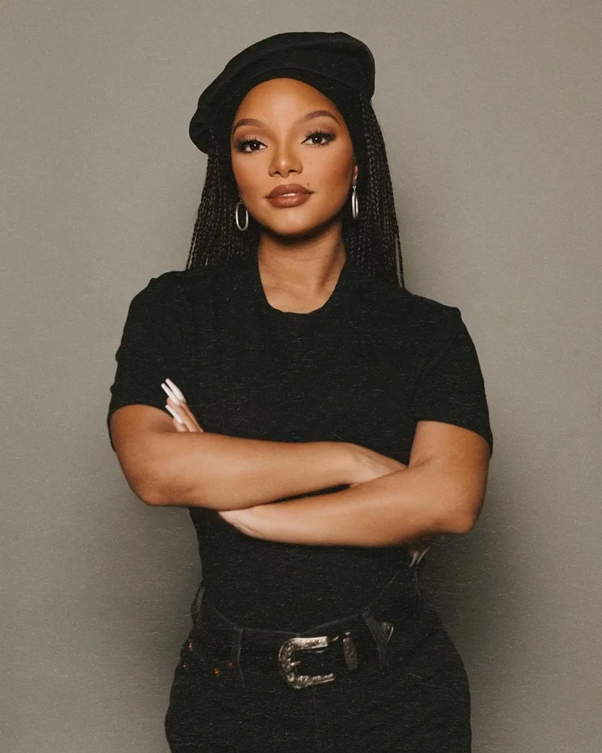 Portrait of a woman with braided hair wearing a beret and a black top, arms crossed, in front of a grey background.