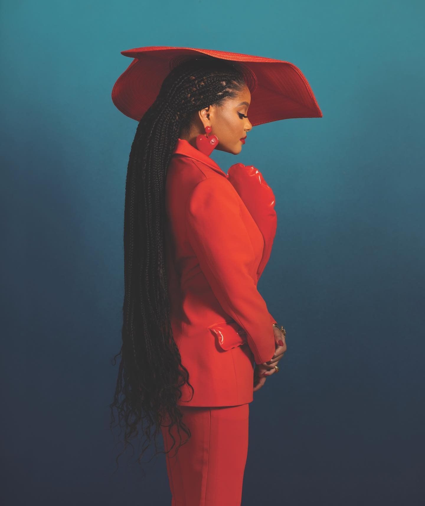 Profile of a woman wearing a red hat and suit against a gradient teal background, showcasing long braided hair and statement earrings.