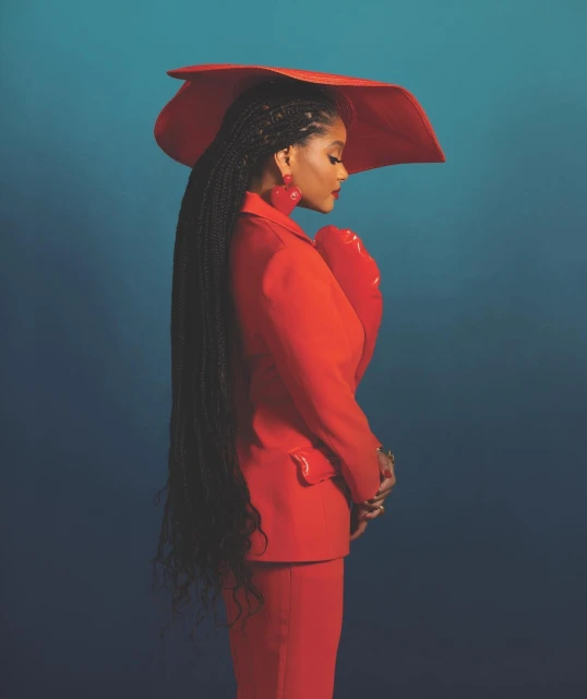 Profile of a woman wearing a red hat and suit against a gradient teal background, showcasing long braided hair and statement earrings.