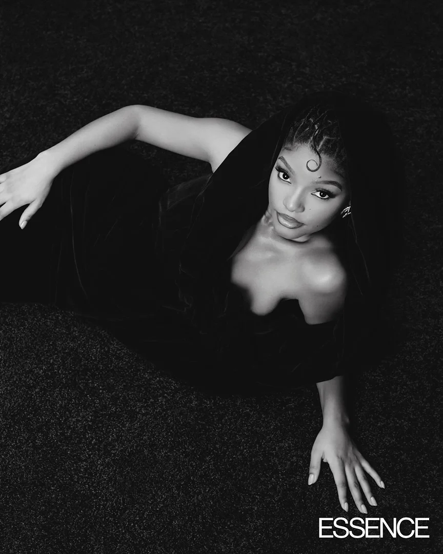 Black and white portrait of a woman with braided hair lying on her side, wearing a black dress against a dark background.