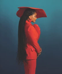 Black woman in red suit and wide-brim hat with braided hair against a gradient blue background