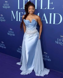 Halle Bailey smiling on a purple carpet in a pale blue strapless gown with crystal detailing and sheer draped overlays.