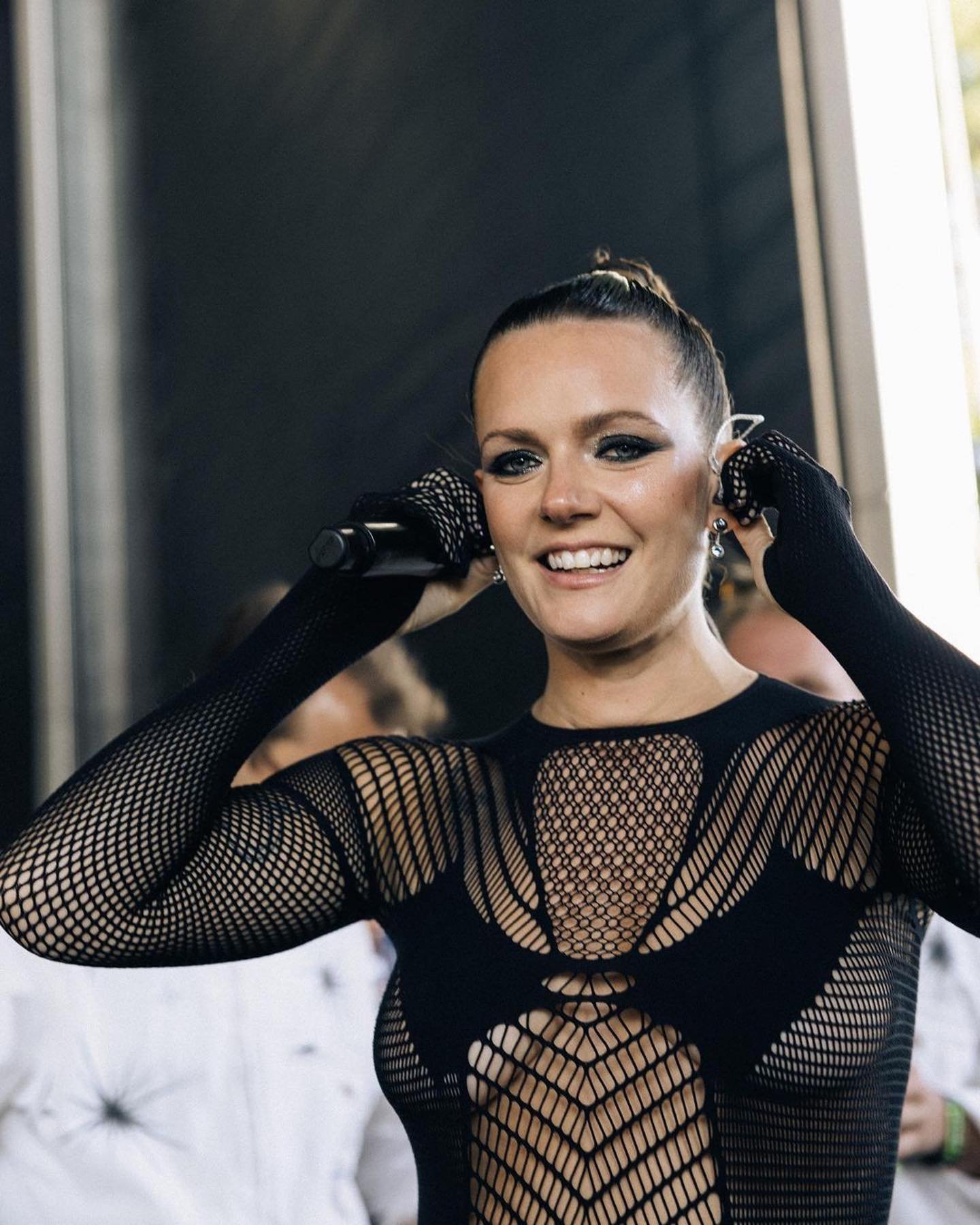 Smiling woman in a black mesh top holds a microphone, likely performing on stage at a music event, with an outdoor setting.