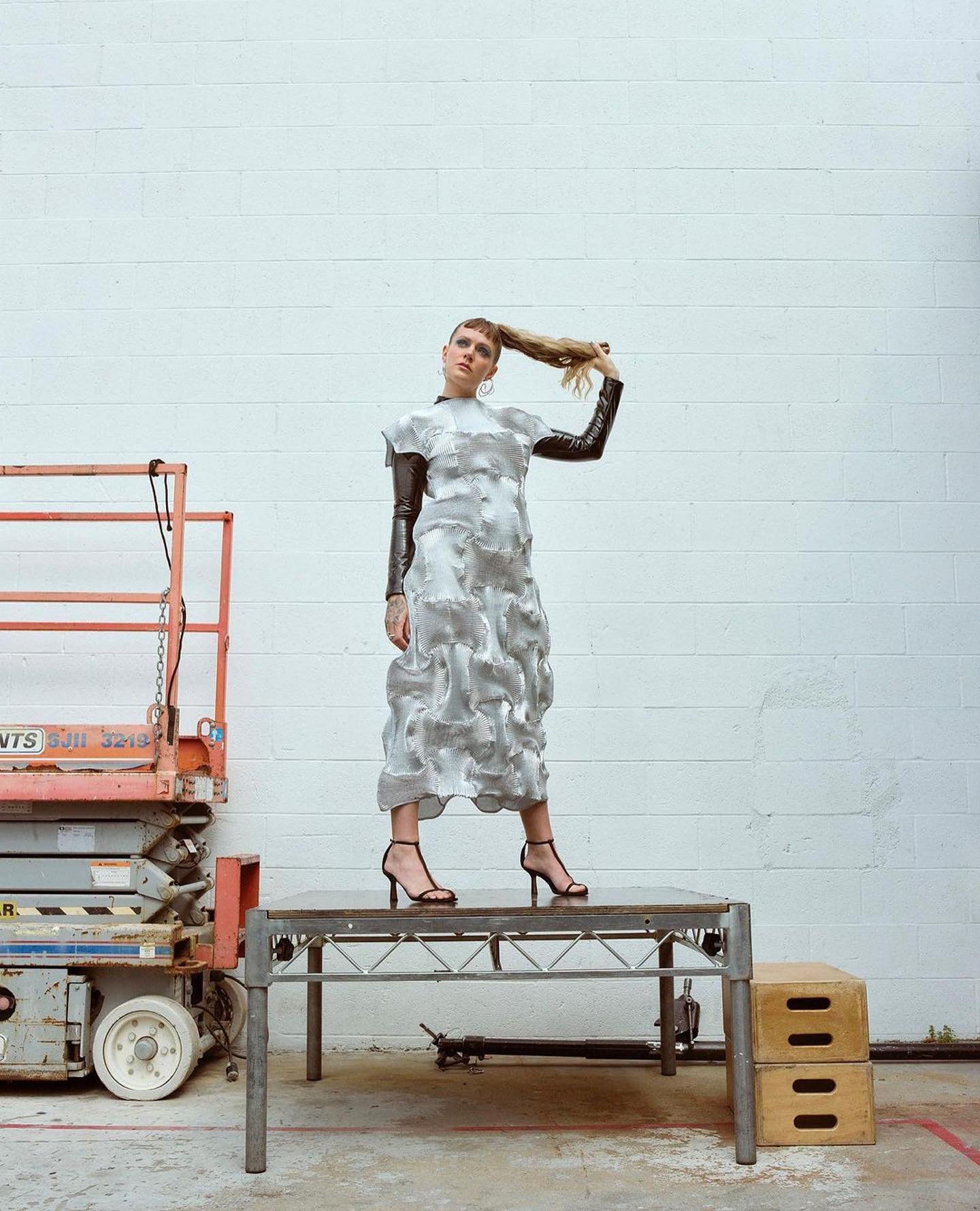 Woman in a silver dress and black sleeves strikes a pose on a table, holding her blonde hair up with a brick wall background and orange lift.