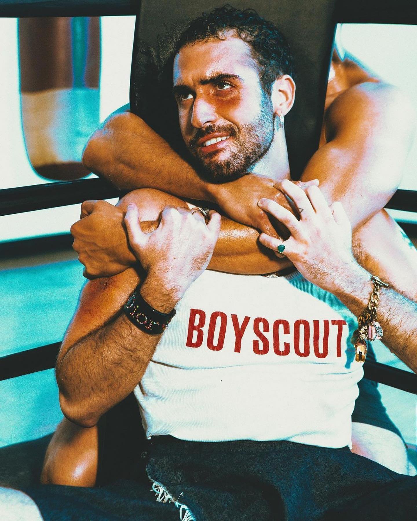 A male boxer being held with a chokehold. He is wearing a white tank top that reads 'BOYSCOUT' with a boxing ring visible in the background.