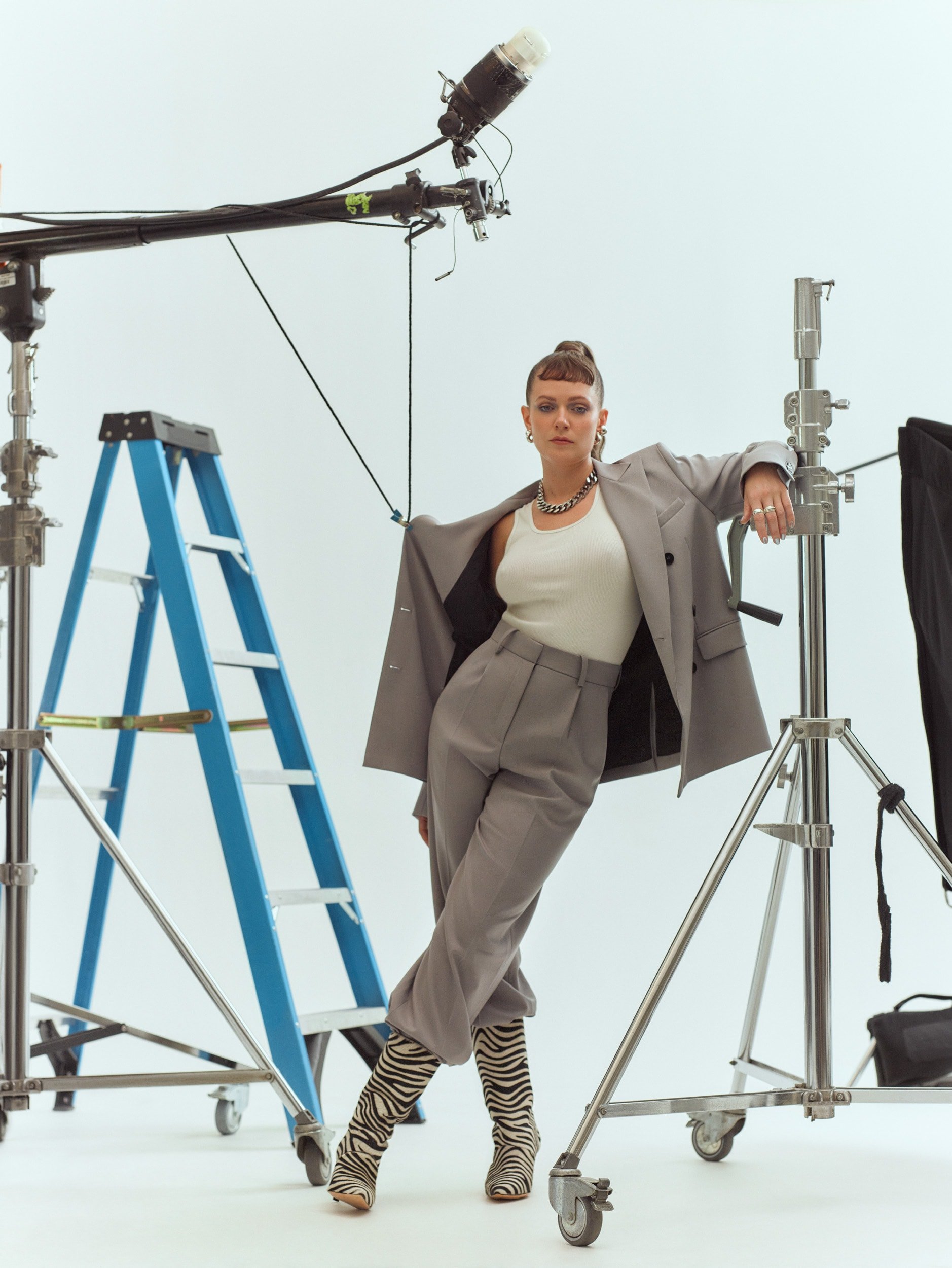 A young woman with a modern hairstyle poses in an open grey suit jacket, pants, zebra print boots and jewelry in a photo studio setting.