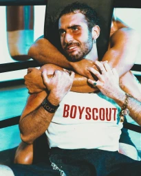 Man wearing a white boyscout tank top held in a headlock by another person in a boxing ring setting.