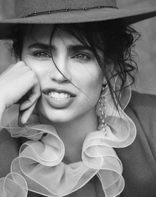 Black and white portrait of a woman wearing a hat and ruffled collar, resting her chin on her hand with a soft, engaging smile.