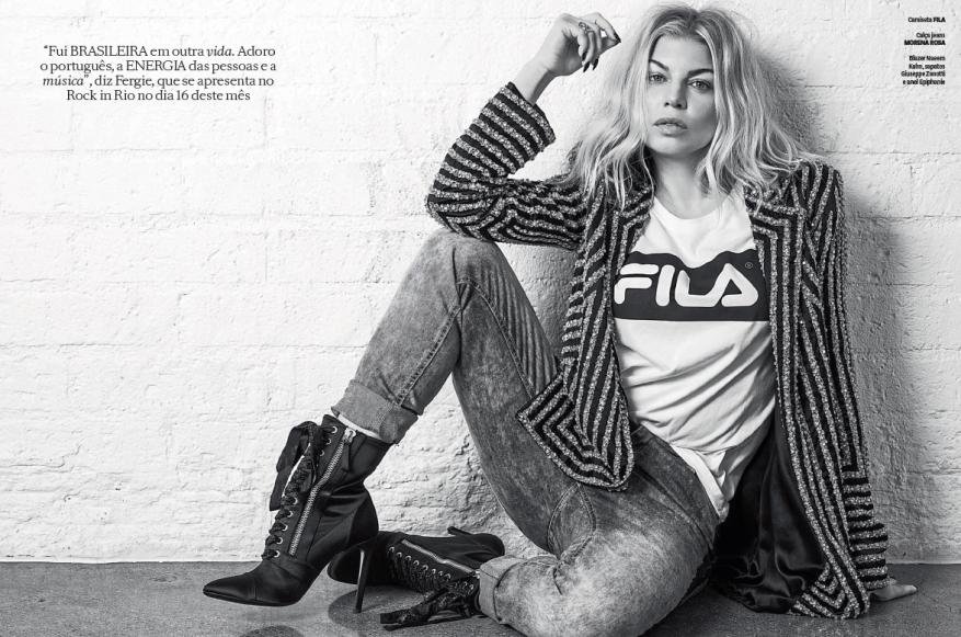 Blonde woman in striped jacket and jeans sits against a white brick wall, wearing a Fila shirt in black and white studio shot.