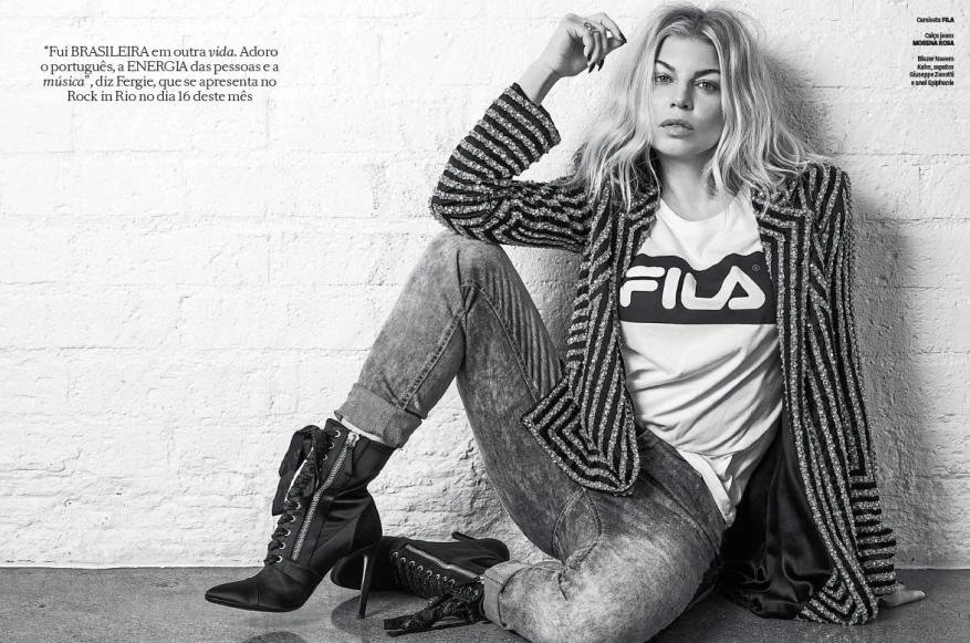 Blonde woman in striped jacket and jeans sits against a white brick wall, wearing a Fila shirt in black and white studio shot.