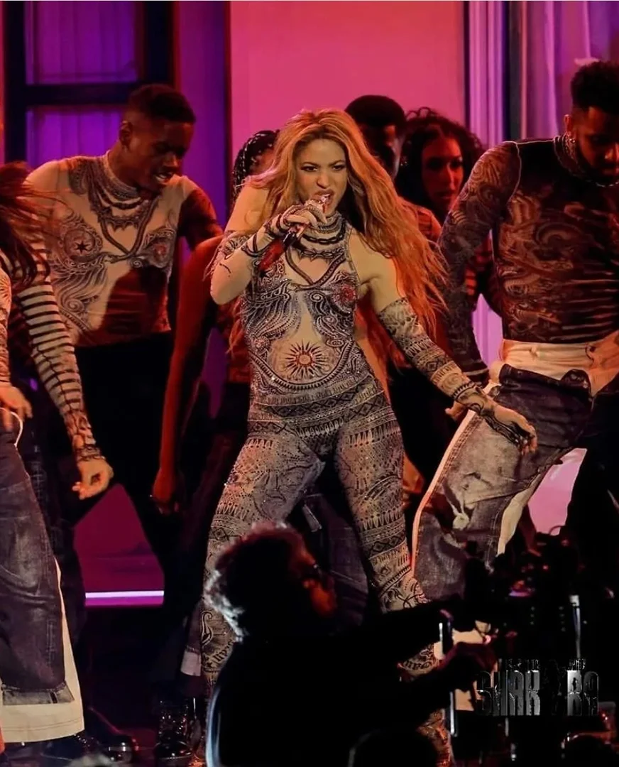 Shakira performs on stage wearing a patterned jumpsuit with dancers in a concert setting with stage lighting and a microphone.