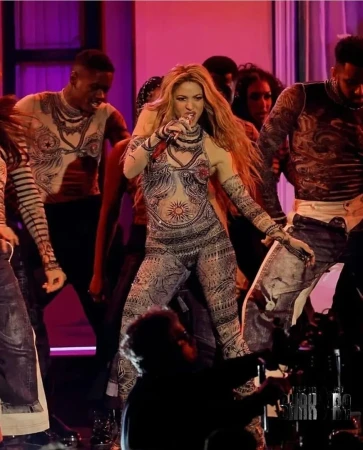 Shakira performs on stage wearing a patterned jumpsuit with dancers in a concert setting with stage lighting and a microphone.