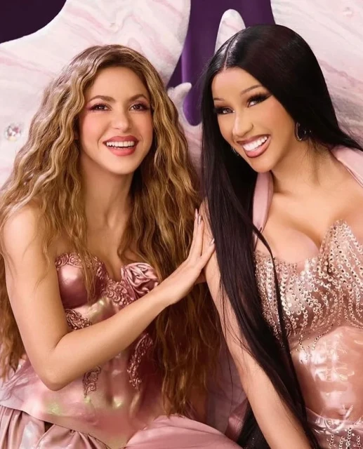 Two women, Shakira and Cardi B, pose in pink corsets with soft lighting. They are smiling in a studio setting with a glamorous feel.