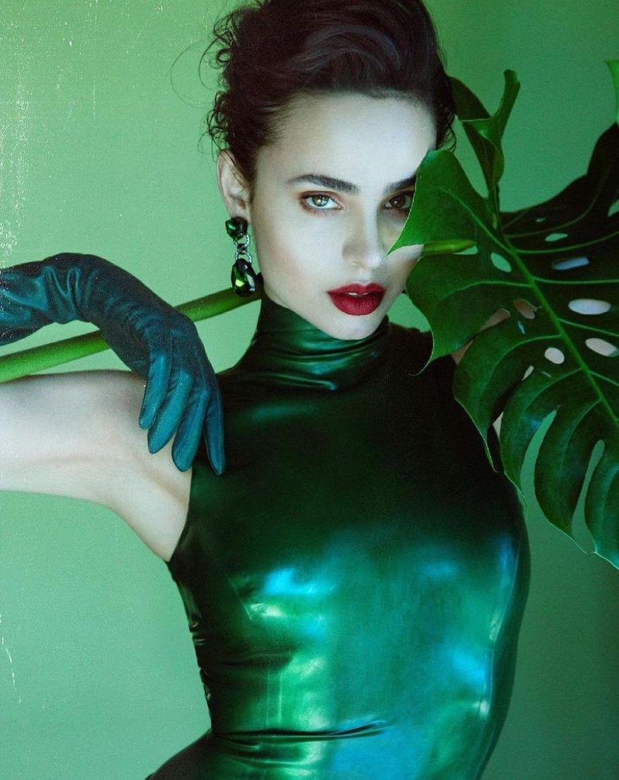 Portrait of an adult woman in a dark green dress, posing in front of a green background with monstera leaves and wearing green earrings.