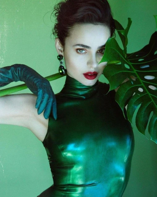 Portrait of an adult woman in a dark green dress, posing in front of a green background with monstera leaves and wearing green earrings.