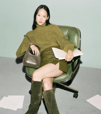 A woman wearing an olive-green turtleneck sweater, skirt, and boots sits in an office chair with a green leather back and seat.