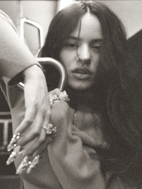 Close-up portrait of a woman with long dark hair, holding onto a metal bar, wearing large rings and decorative nails in a photo studio setting.
