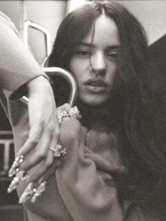 Close-up portrait of a woman with long dark hair, holding onto a metal bar, wearing large rings and decorative nails in a photo studio setting.