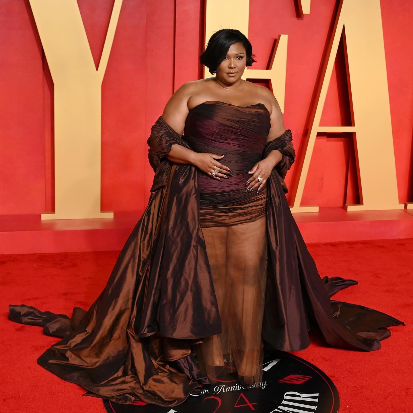 Lizzo poses on a red carpet wearing a brown strapless dress with a long train and matching shawl, smiling at the camera.