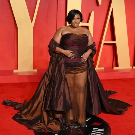 Lizzo poses on a red carpet wearing a brown strapless dress with a long train and matching shawl, smiling at the camera.