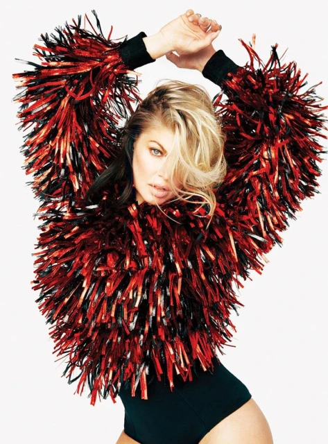 Portrait of a blonde woman with hands above her head wearing a black bodysuit and red and black tinsel jacket on a white background.