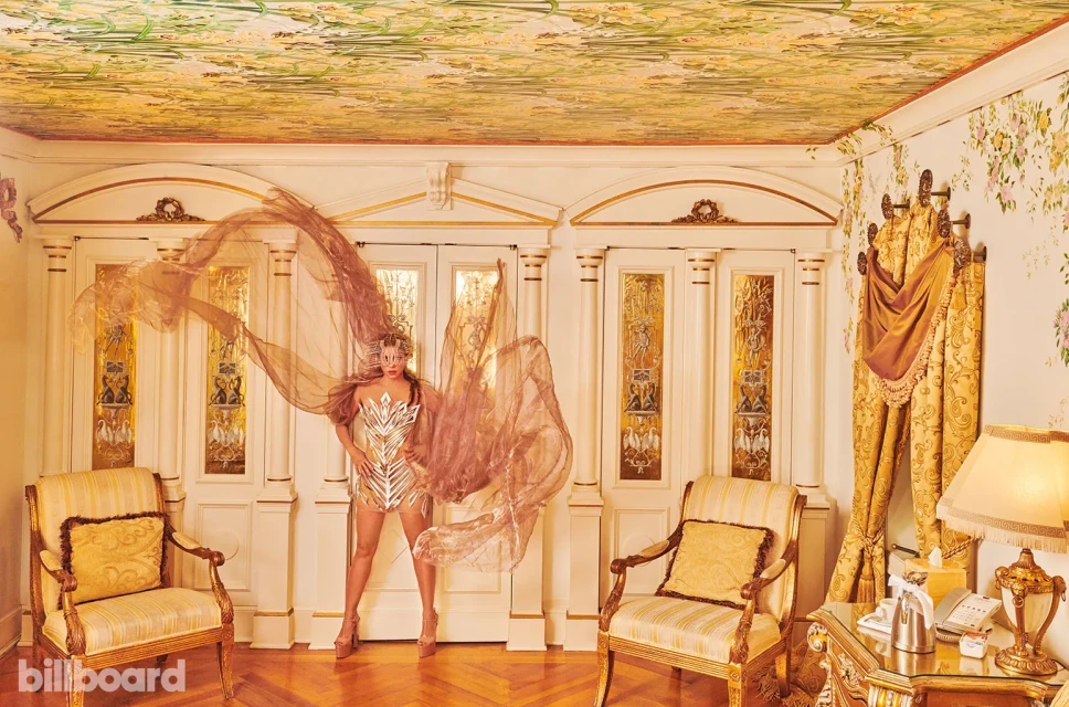 Woman in a metallic dress poses indoors with a flowing chiffon fabric, ornate doors, floral wallpaper, and gilded accents.