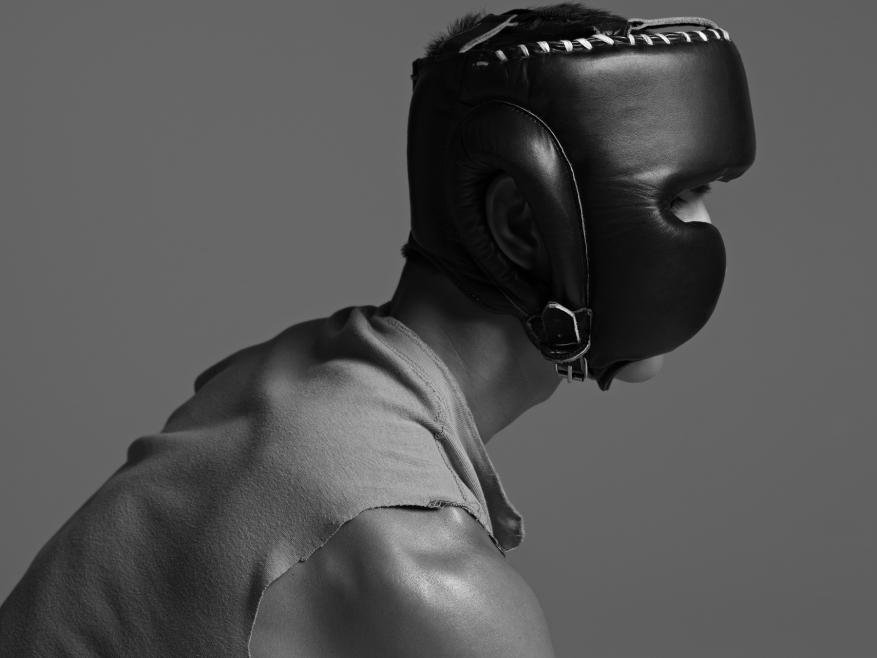 Monochromatic portrait of an adult male wearing a black leather boxing headgear, looking away in a simple studio setting.