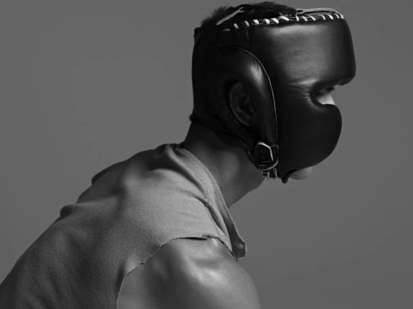 Monochromatic portrait of an adult male wearing a black leather boxing headgear, looking away in a simple studio setting.