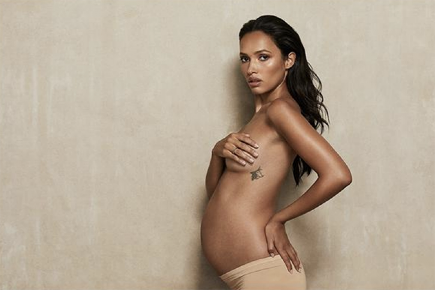 Portrait of a pregnant woman in nude underwear holding her chest with a tattoo, indoors against a light tan background.