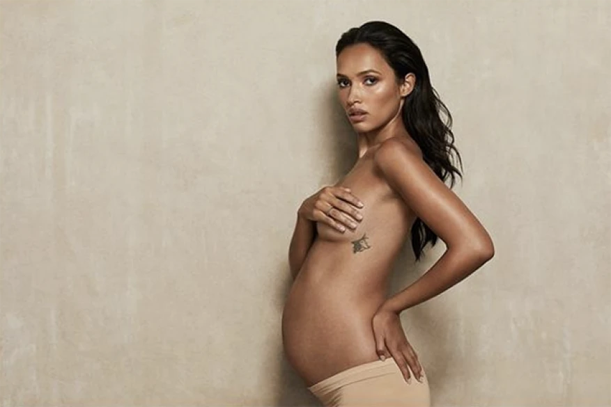 Portrait of a pregnant woman in nude underwear holding her chest with a tattoo, indoors against a light tan background.