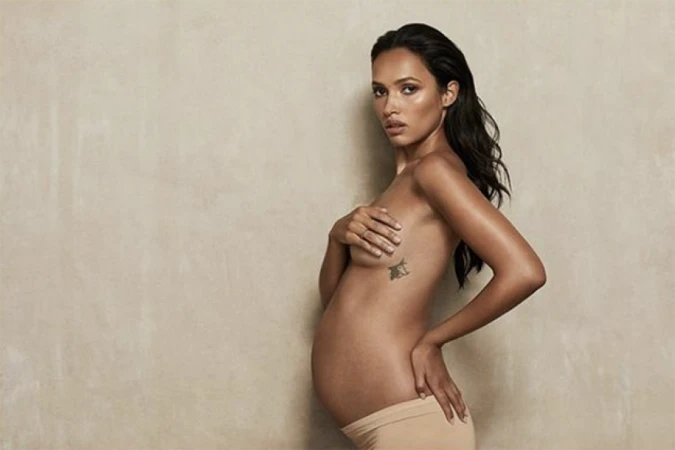 Portrait of a pregnant woman in nude underwear holding her chest with a tattoo, indoors against a light tan background.