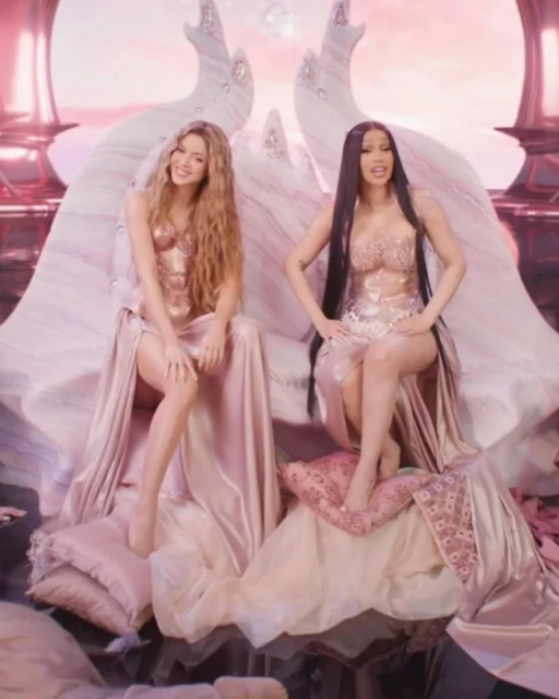 Two adult females, Shakira and Cardi B, sit on an ornate throne made of pillows in a digitally enhanced, fantastical setting with pink lighting.