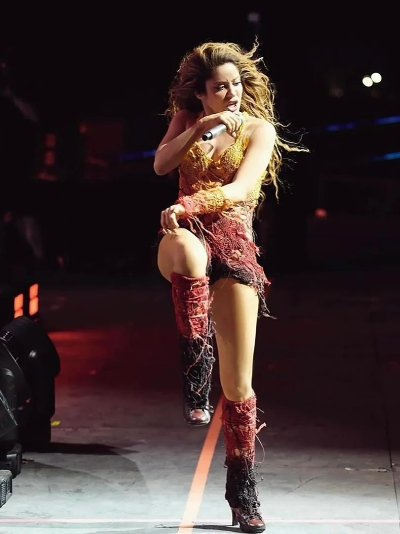 A woman performs on stage wearing a yellow and red fringed outfit, high-heeled boots, and holding a microphone while kicking one leg up.