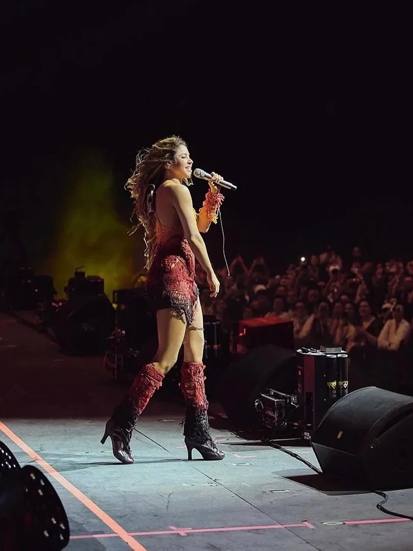 Singer performs on stage in a red dress and high heels with a microphone, backed by a crowd and concert lighting at a live music event.