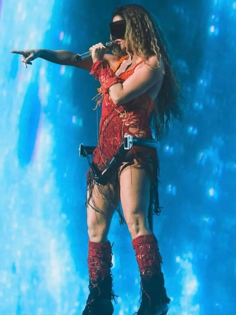 A person in a red, fringed outfit stands on a stage with a microphone, pointing to the left against a backdrop of blue lights.