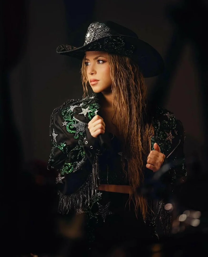 Portrait of a blonde woman in a black, star-studded cowboy hat and jacket, glancing left in a dimly lit setting, exuding a country-western vibe.