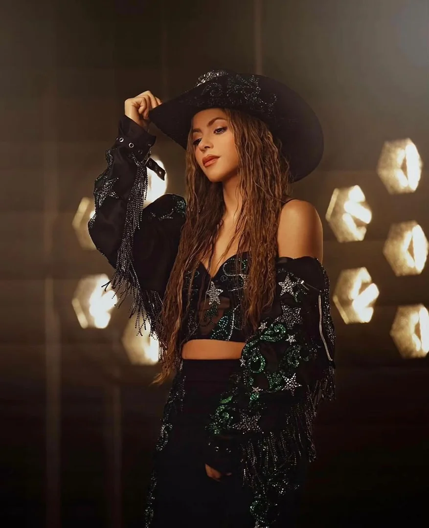 Portrait of Shakira wearing a wide-brimmed black hat and a sequined, sheer black top and skirt. The background features out-of-focus lights.
