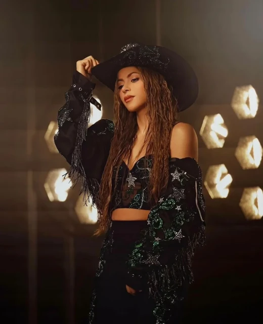 Portrait of Shakira wearing a wide-brimmed black hat and a sequined, sheer black top and skirt. The background features out-of-focus lights.