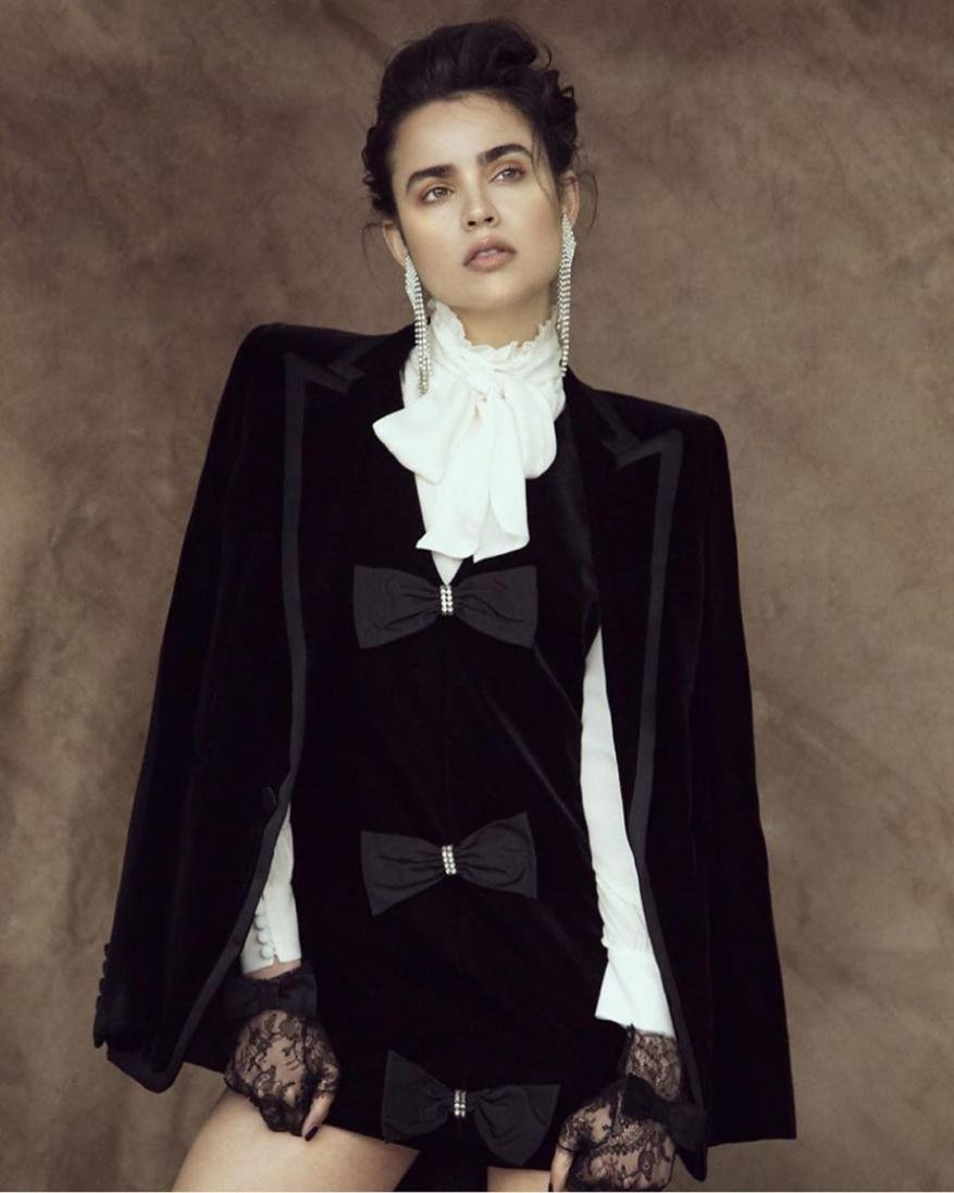 A woman in a black velvet dress with bow details and a white ruffled collar stands in a studio, looking up with dangling earrings.