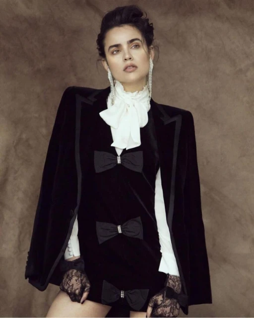 A woman in a black velvet dress with bow details and a white ruffled collar stands in a studio, looking up with dangling earrings.