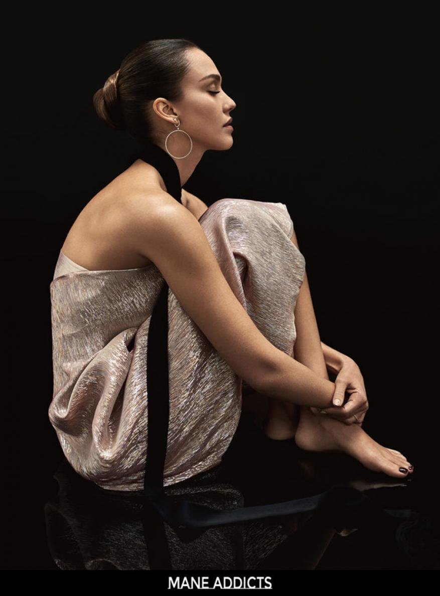 Portrait of Jessica Alba in elegant dress sitting in photo studio with dark backdrop, wearing hoop earrings and looking serene.