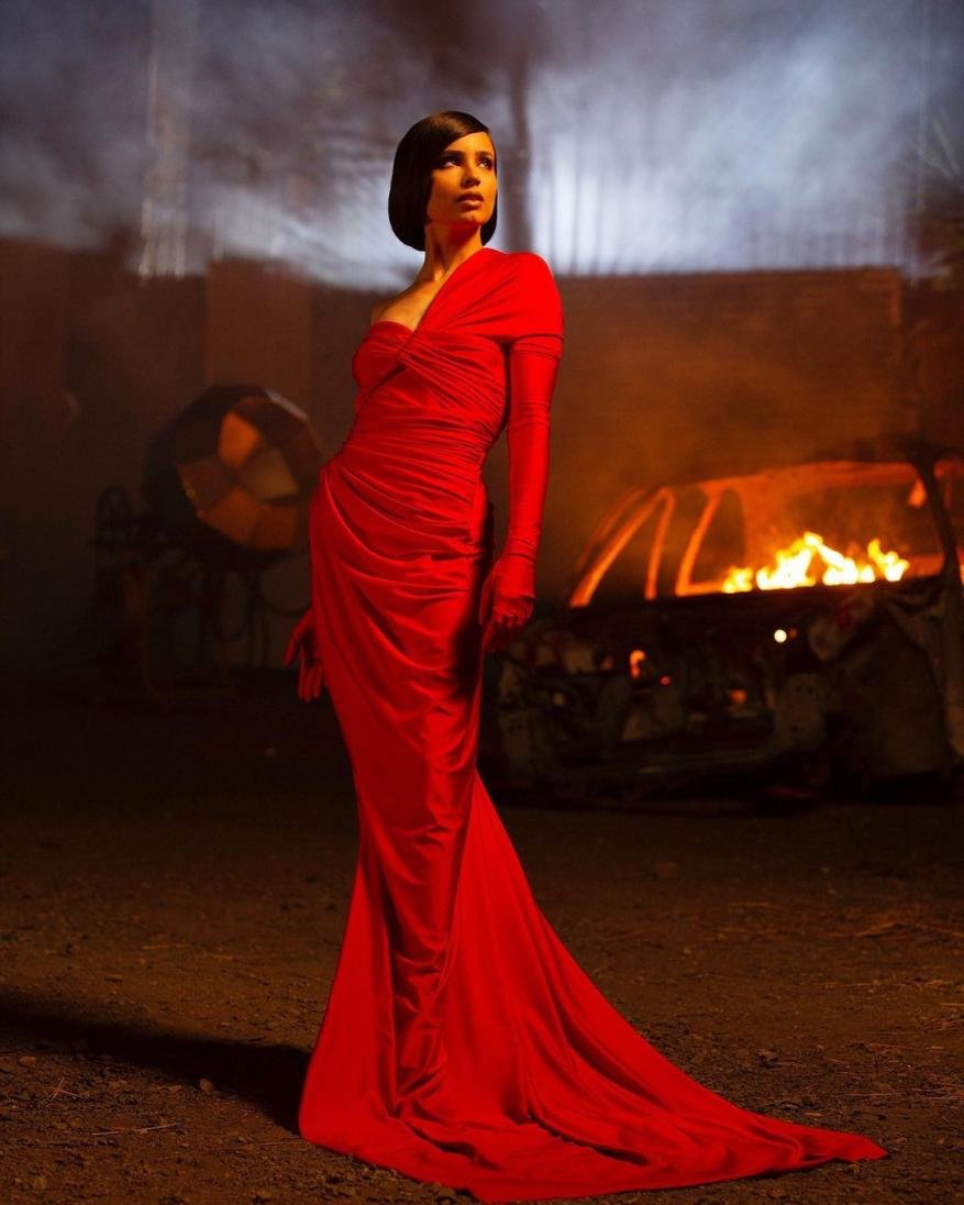 Elegant woman in a red gown stands near a burning car against a dimly lit, apocalyptic background, creating a striking contrast.